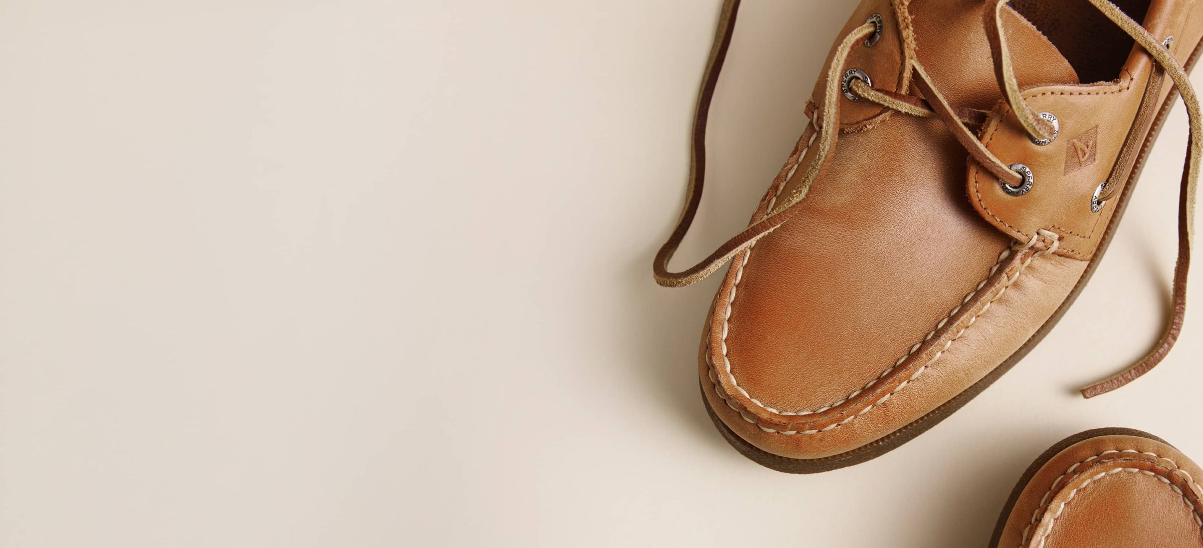A close-up of a tan Sperry Authentic Original leather boat shoe with matching laces, photographed on a clean off-white background with a second shoe partially visible at the bottom right.
