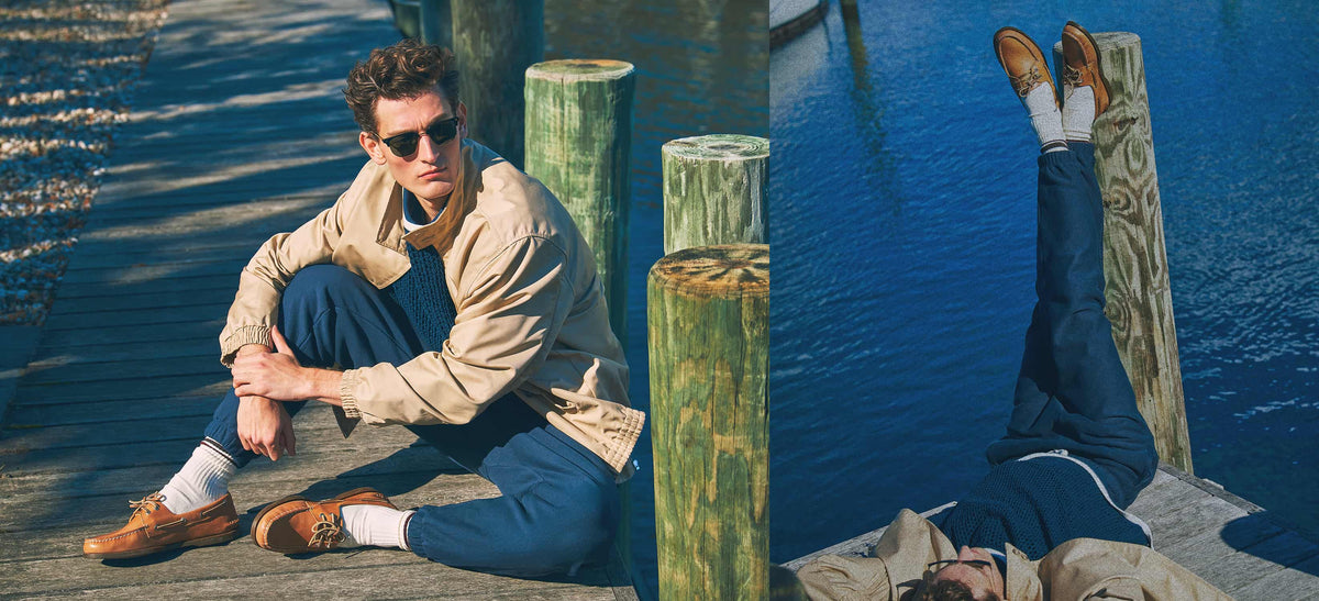 A man wearing a beige jacket, navy pants, white socks, and tan leather Sperry boat shoes sits on a wooden dock next to water, with a second view showing his legs kicked up against a post.